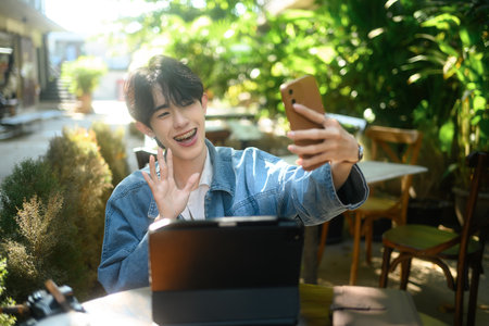 Smiling young asian man with braces waving at the camera while video calling at outdoor cafe.の写真素材