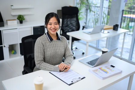 Cheerful businesswoman sitting at her desk in a modern office, smiling at the camera.の写真素材