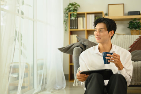 Young asian man holding a coffee cup and looking out the window.の写真素材