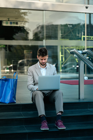 Professional businessman sitting on the steps outside a modern glass building, working on his laptop.の写真素材