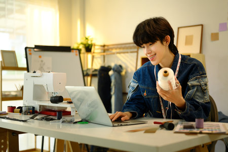 Smiling designer holding a spool of thread working on laptop in a stylish workshop.