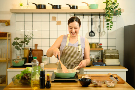 Smiling middle aged woman stirring ingredients in frying pan while preparing meal in cozy kitchen.の写真素材
