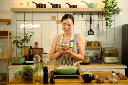 Smiling adult woman checking recipe on smartphone while cooking in kitchen.の写真素材