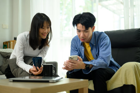 Young couple relaxing in living room and sharing social media news on mobile phone.の写真素材