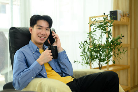 Smiling man enjoying a coffee and talking on the phone in a cozy home.の写真素材