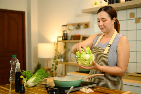 40s asian woman adding fresh vegetables to a pan while cooking in a kitchen.の写真素材