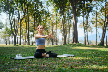 Beautiful woman sitting on yoga mat in a park stretching her arms and enjoying the fresh air.