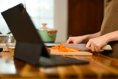 Young woman chopping carrots for spaghetti sauce and following a recipe on digital tablet in a kitchen.の写真素材