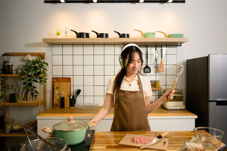Cheerful asian woman cooking in a cozy kitchen and listening to music with headphones.の写真素材