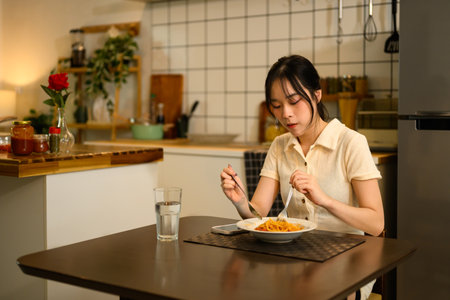 Pretty asian woman sitting at a dining table, enjoying a cozy meal in a warm kitchen.の写真素材