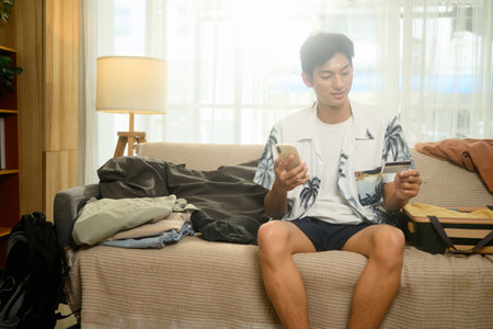 Young man in summer clothes holding smartphone and credit card sitting on couch with packed luggage.の写真素材