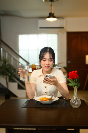 Happy young woman sitting at dining table with a plate of spaghetti and checking mobile phone.の写真素材