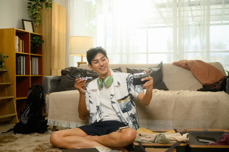 Cheerful man holding vintage camera and toy airplane while packing suitcase, excited for his...