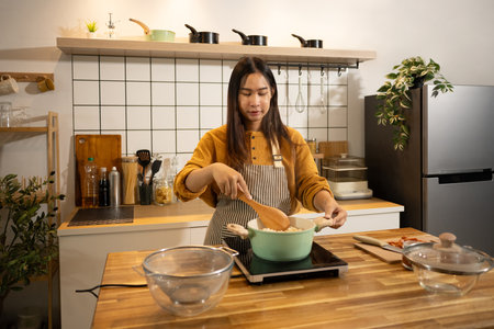 Young asian woman stirs pot on stove, preparing a homemade meal in a kitchen.の写真素材