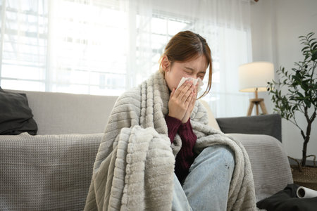 Young woman suffering from flu symptoms wrapped in blanket, sneezing into a tissue..の写真素材