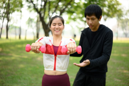 Smiling woman lifting light dumbbells with trainer guidance in a green park.の写真素材