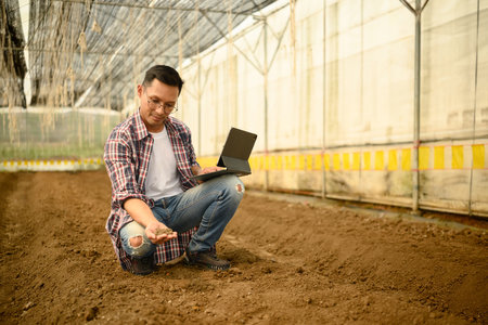 Male agronomist holding digital tablet and assessing soil conditions in a prepared greenhouse plot.の写真素材