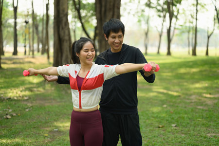 Man assisting woman with dumbbell exercise in a sunny park.の写真素材