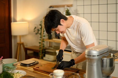 Smiling young man chopping fresh parsley on wooden cutting board while preparing garlic butter in a cozy kitchen.の写真素材