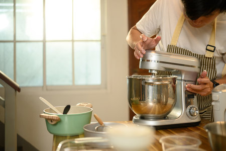 Young asian man operating a stand mixer to whip cream in a cozy kitchen.の写真素材
