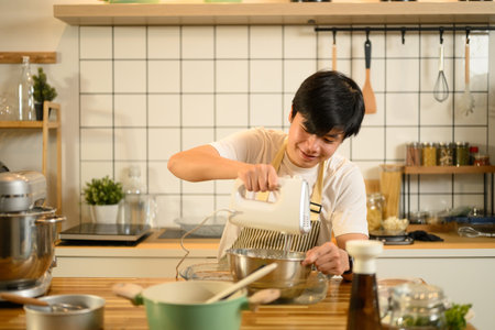 Smiling asian man using handheld electric mixer to whip cream in a stainless steel bowl.