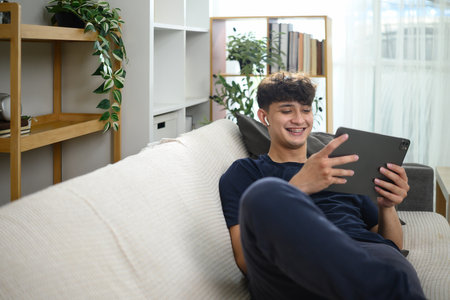 Smiling man relaxing on sofa watching content on digital tablet with wireless earbuds.の写真素材