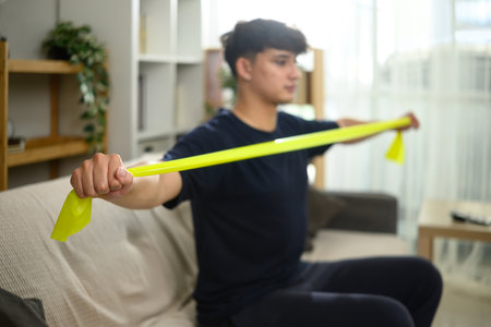Young man exercising with yellow resistance band while sitting on sofa at home.