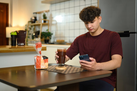 Young man sitting at kitchen table drinking chocolate protein shake and using smartphone.