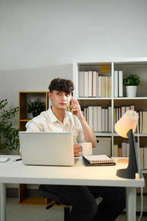 Caucasian young entrepreneur using his laptop while talking on mobile phone at workplace.の写真素材