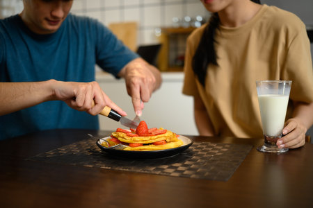 Happy young couple enjoying pancakes with strawberries and syrup in cozy home kitchen.の写真素材