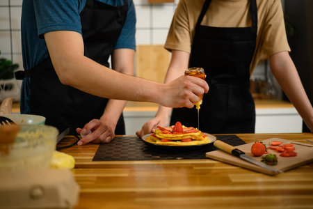 Close-up of man pouring honey over stacked pancakes with strawberries in a cozy kitchen..の写真素材