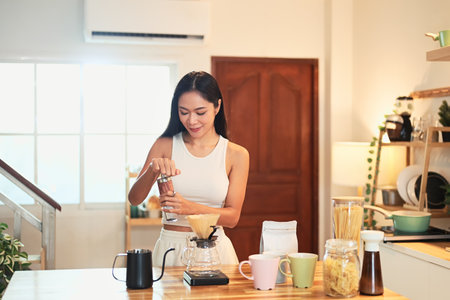 Young woman grinding coffee beans and preparing pour over coffee in a warm kitchen.の写真素材