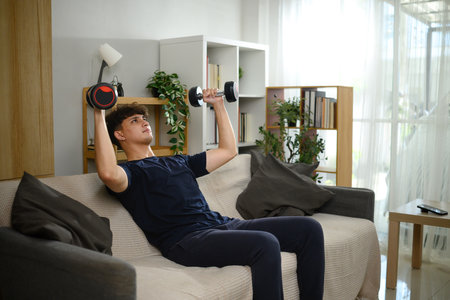 Young man lifting dumbbells on a sofa. Fitness at home concept.の写真素材