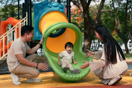 Happy parents playing with their toddler on a colorful playground slide at the park.の写真素材
