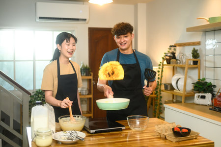 Cheerful couple cooking breakfast in a cozy kitchen, flipping pancakes with smiles and laughter.の写真素材