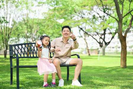 Father and daughter enjoy a sunny day in the park sitting together on a bench.の写真素材