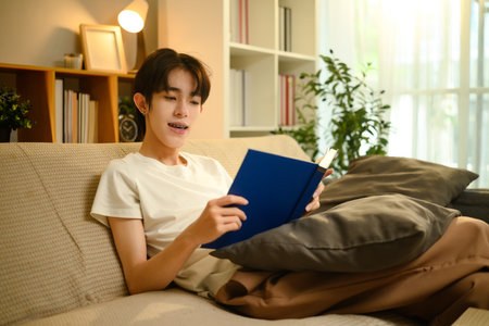 Happy man reading a book while relaxing on sofa in a bright living room.の写真素材