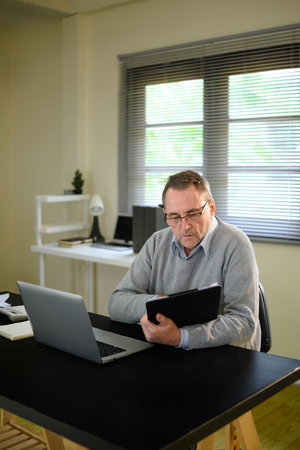 Focused senior man in glasses works at home with digital tablet and laptop.の写真素材