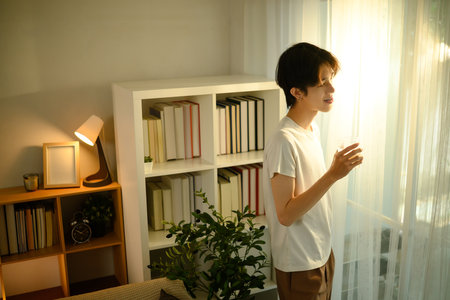 Young man standing by the window, enjoying the morning sunlight in a cozy home.の写真素材