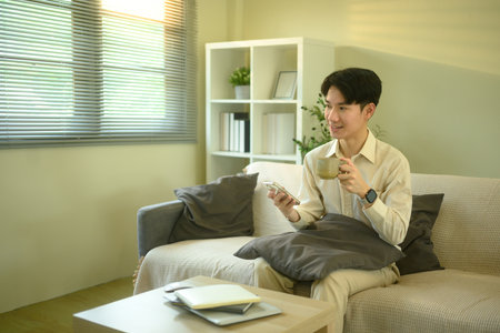 Smiling man checks messages or notifications on smartphone while seated in living room.の写真素材