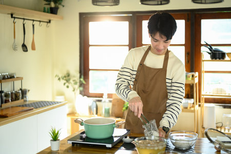 Smiling man in an apron prepares noodles over a stove in warm modern kitchen.の写真素材
