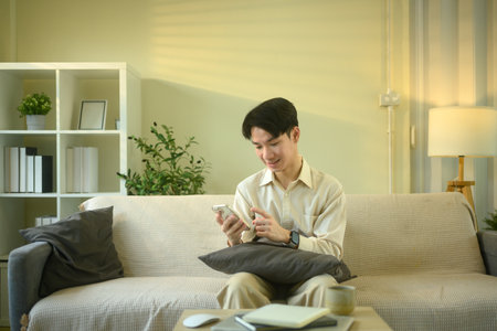 Smiling man checks messages or notifications on smartphone while seated in living room.の写真素材
