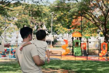 Father carries his toddler while walking toward a vibrant play area in a park.の写真素材