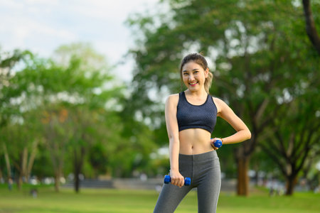 Portrait of a cheerful female athlete lifting dumbbells in a green park.の写真素材