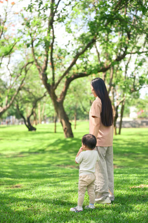 Asian mother and her toddler stroll through a lush green park.の写真素材