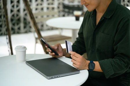 Man holding credit card and using smartphone for an online transaction at an outdoor cafe.の写真素材