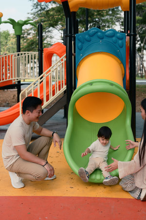 Happy parents watch and cheer as their toddler slides down a colorful slide.の写真素材
