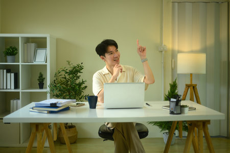 Smiling man entrepreneur sitting at working desk and pointing finger up.の写真素材