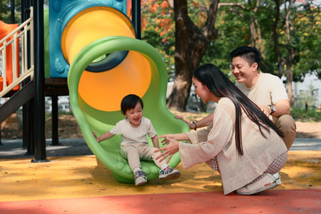 Happy parents watch and cheer as their toddler slides down a colorful slide.の写真素材