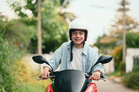 Cheerful young man wearing helmet riding a red motorcycle on a quiet residential street.の写真素材